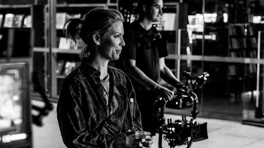 A woman in a ponytail smiles confidently on a film set as a cameraman behind her moves a camera rig along the floor.