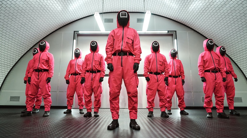 A still from the Netflix show Squid Game, with a group of people in pink hazmat suits and black masks