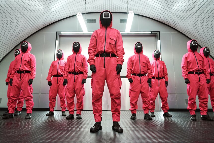 A still from the Netflix show Squid Game, with a group of people in pink hazmat suits and black masks