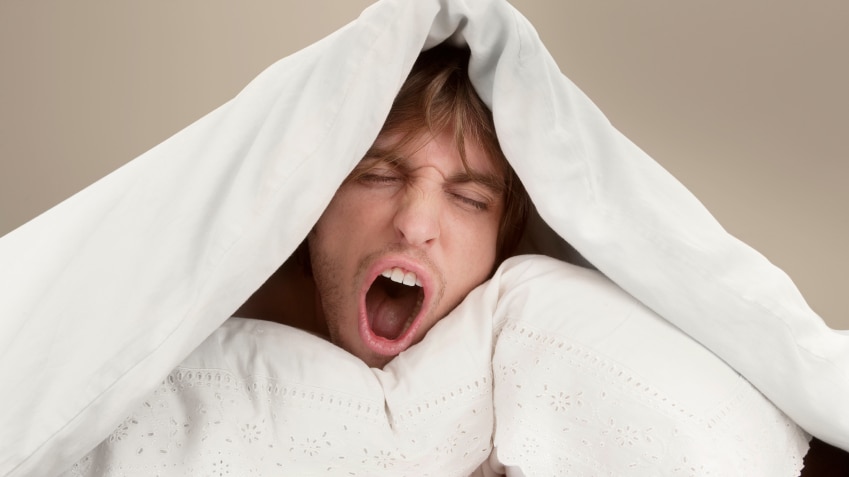 Young man with pillow under quilt yawning