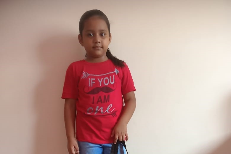 A young girl wearing a red tshirt standing against a wall.