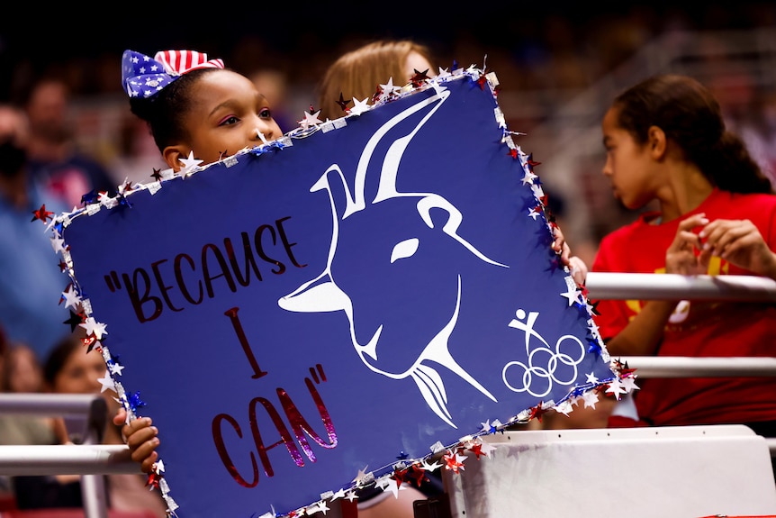 A little girl with a USA-print bow in her hair holds a sign that says 'because i can' with a picture of a goat on it in a crowd