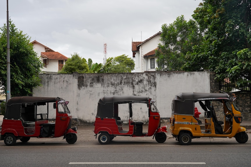 Three auto-rickshaws, two red and one yellow, stand stationary on a road in front of a stained brick wall