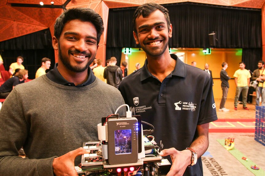 Two male university students stand holding their robot during a robotics competition