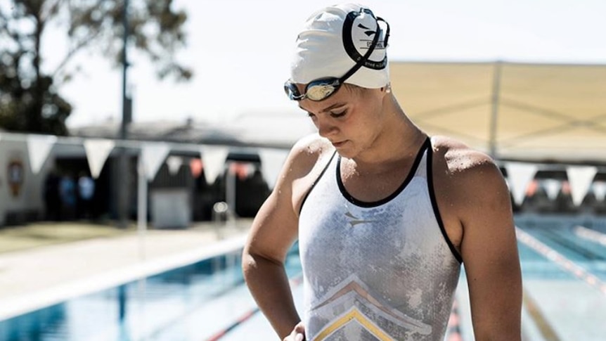 A female swimmer wearing a cap and goggles above her eyes stands beside a swmming pool