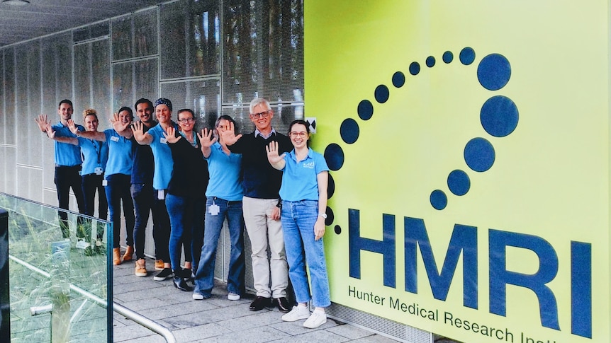 Nine people line up in front of an HMRI sign, each with one arm held out showing five fingers and 
