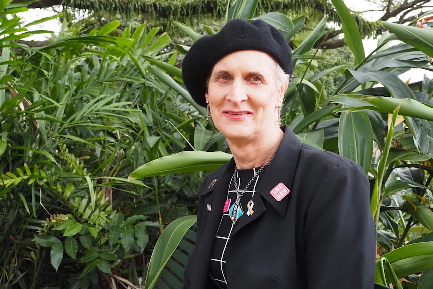 A woman in black coat, dress and beret sits in a tropical garden