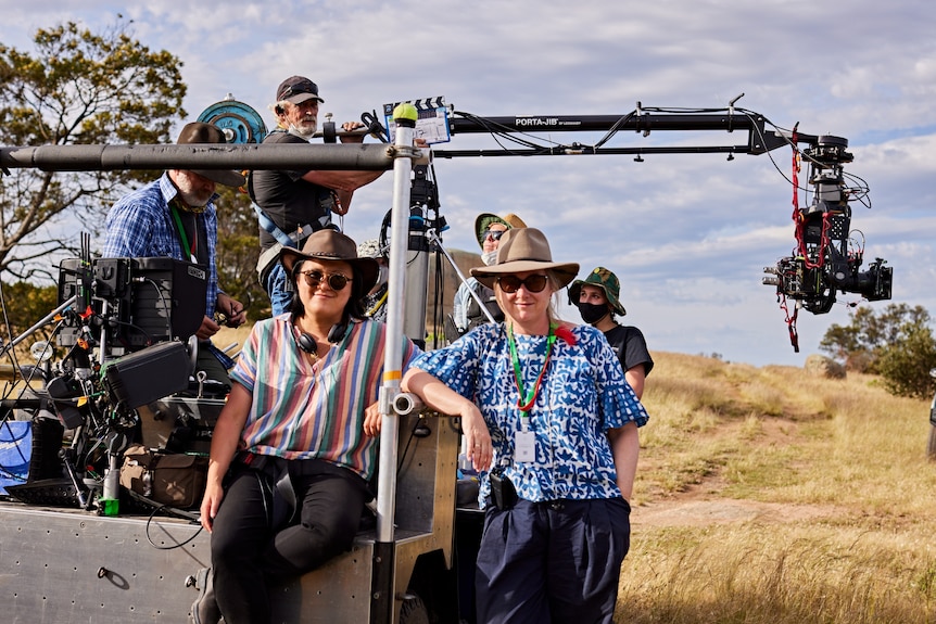 A Taiwanese woman and a white woman both in hates and sunglasses lean on a camera rig with crew, dry bushland in the background