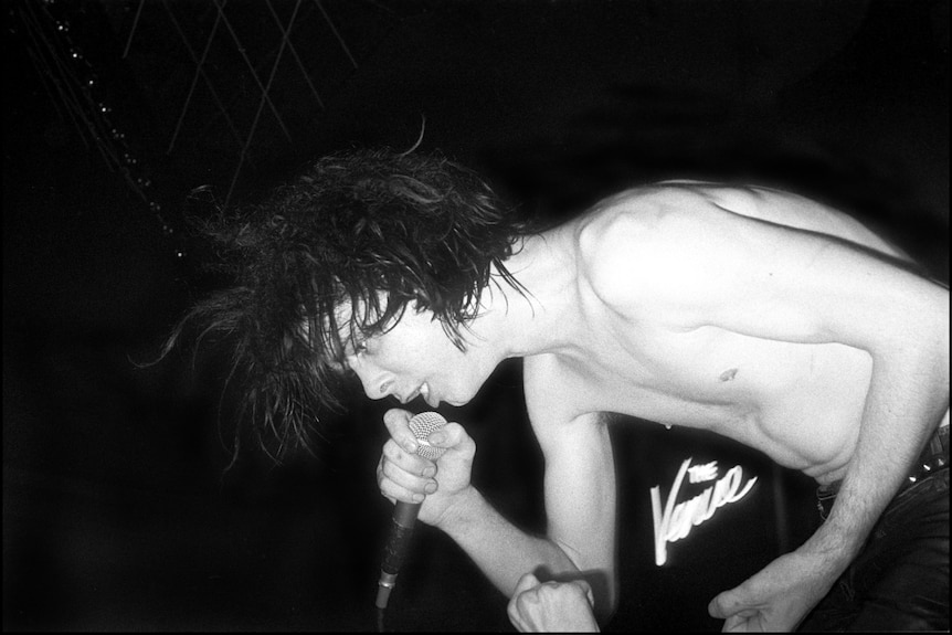 Bare-chested man leans over and sings into a microphone. 