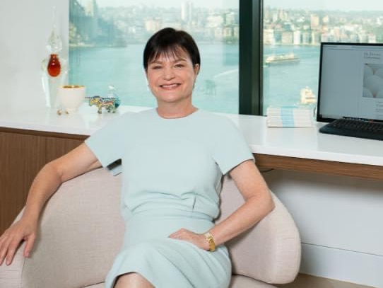 A doctor with short dark hair smiles at the camera while sitting in a chair in an office overlooking the water.
