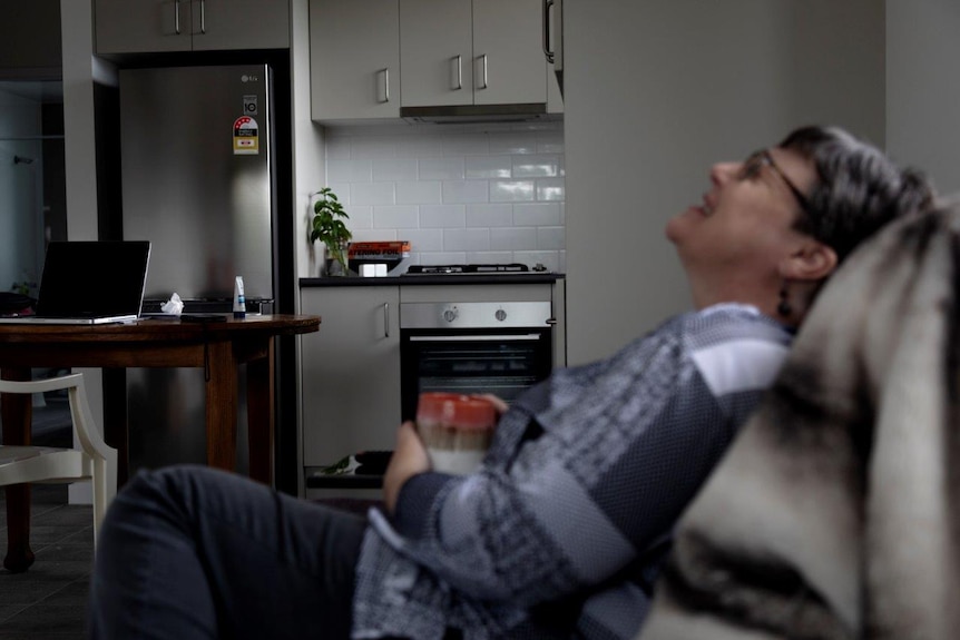 A woman with short brown hair and glasses, and a grey patterned shirt sits of a couch drinking coffee and laughs.