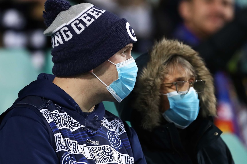 A young man and and older woman wear surgical face masks as they sit together in a sports stadium.