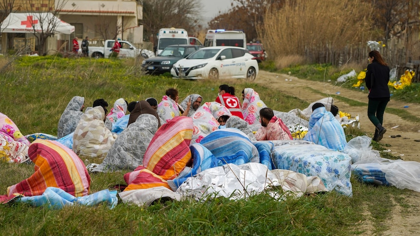 18 people huddle in blankets on the grass to get warm after swimming to shore.