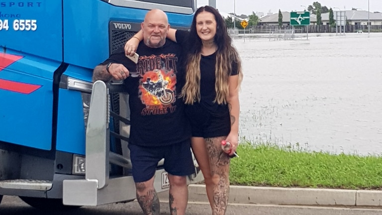 Male truck driver and woman stand in front of large truck with flooded road in the background. They are safe and smiling.