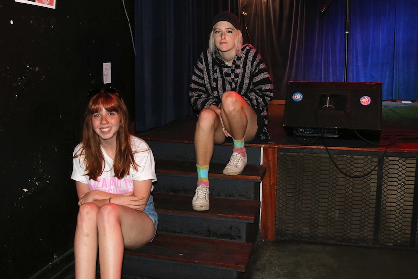Two young women sit on some steps leading up to a stage.