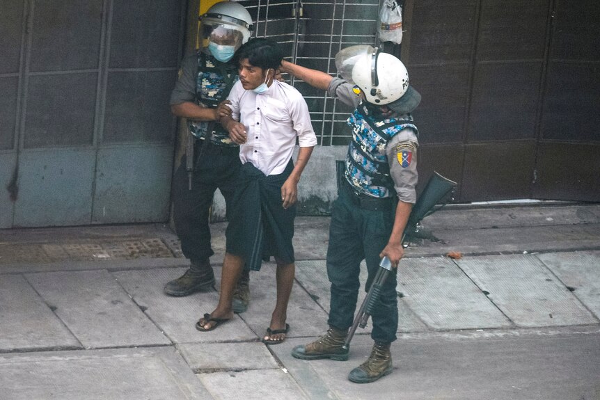 A small Asian man in a white shirt is held by two police officers wearing helmets.