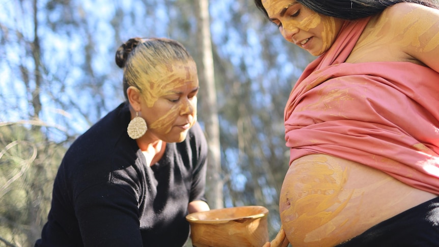 The pregnant belly of a an Indigenous woman is painted by another Indigenous woman with paint on her face