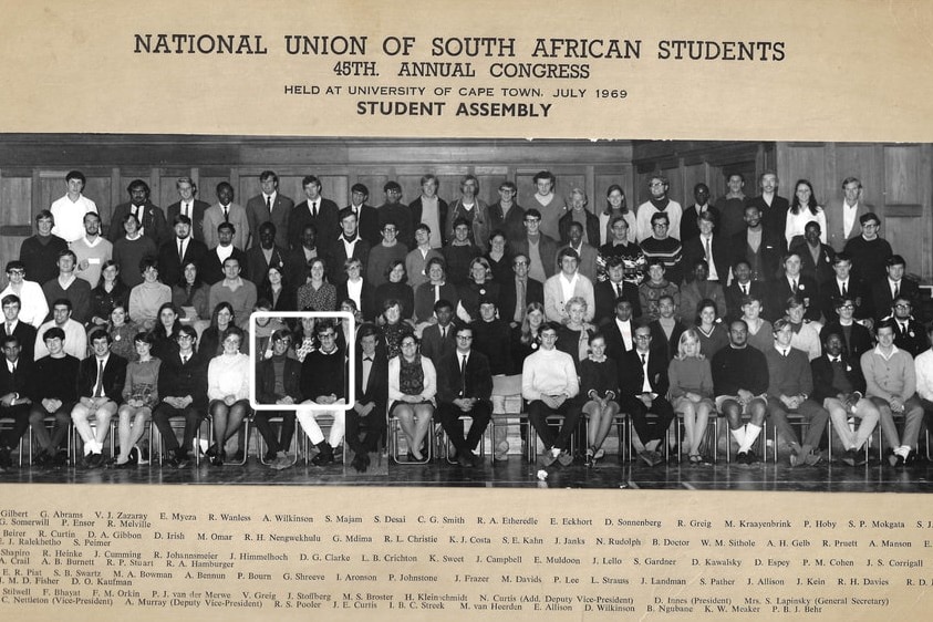 Students pose for school-photograph style shoot, legs crossed and hands on knees. First three rows are seated, back three stand