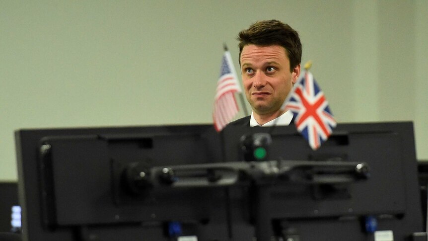 A man in his mid 30s stands in front of a computer embellished with american and union jack flags looking gobsmacked.