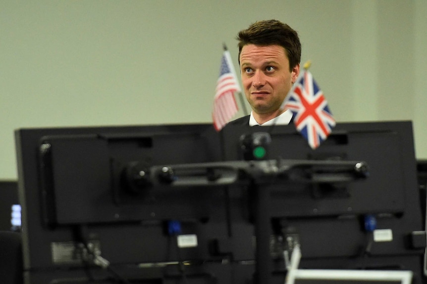 A man in his mid 30s stands in front of a computer embellished with american and union jack flags looking gobsmacked.