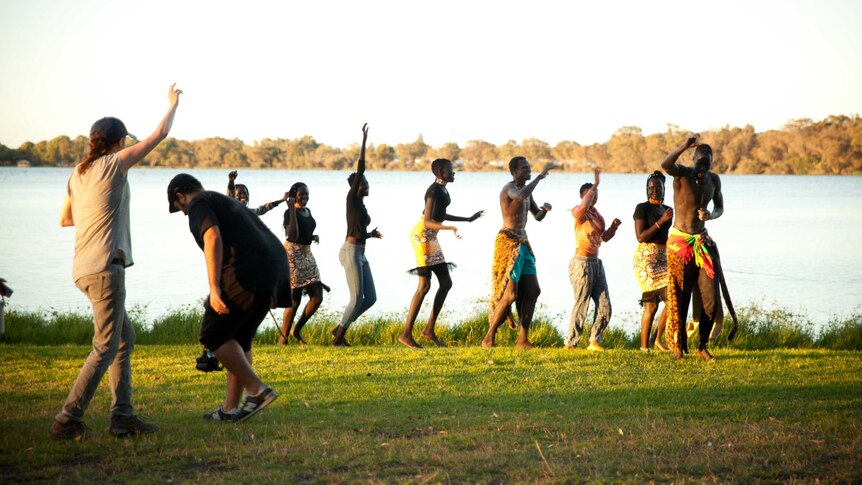 A line of eight dancers at Lake Monger perform for a camera crew wearing African-inspired clothing.