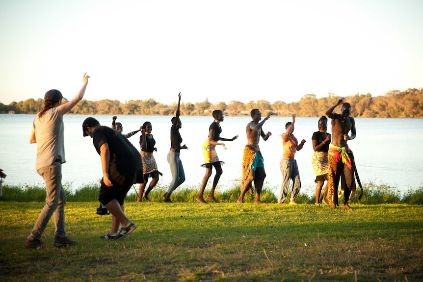 A line of eight dancers at Lake Monger perform for a camera crew wearing African-inspired clothing.