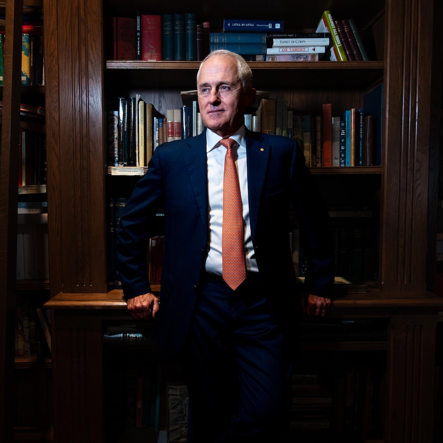 Dressed in a dark suit, white shirt and orange tie, Malcolm Turnbull stands by a bookcase, looking to the side.