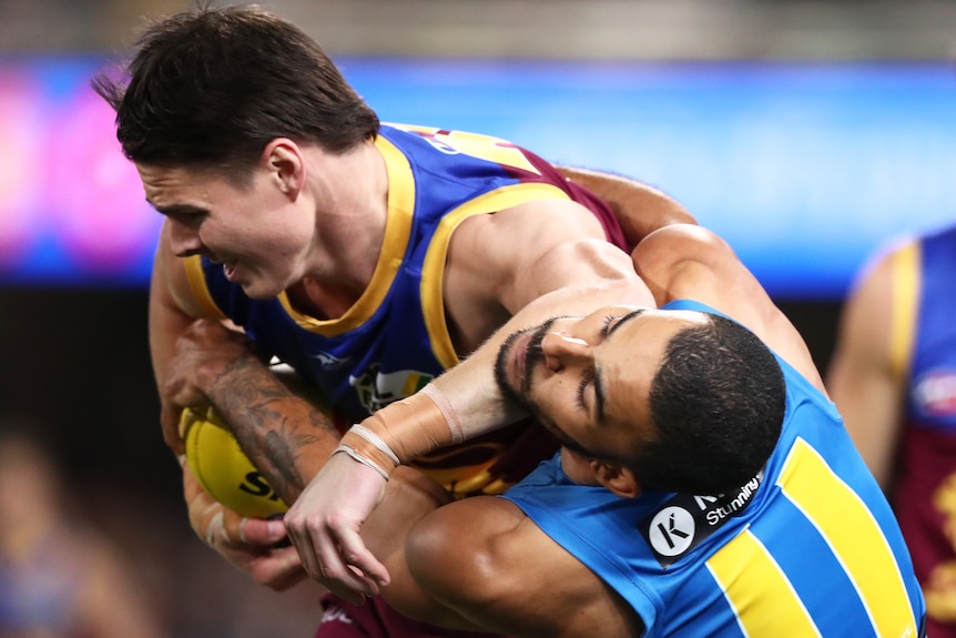 A Brisbane Lions AFL player holds the ball as he fends off a Gold Coast Suns opponent.