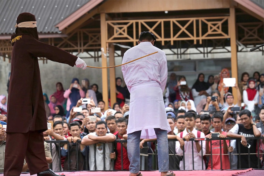 A sharia law official standing on a stage whips a man on the back as a crowd watches on.