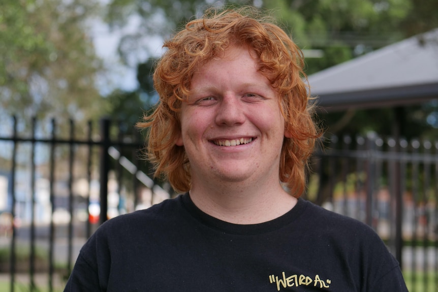 A tall, 15 year old boy stands smiling at the camera. He has red hair and is wearing a black t-shirt.
