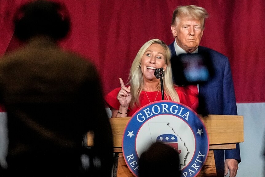 A blonde woman in red speaks animatedly on stage in front of sombre Donald Trump on stage
