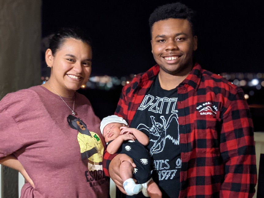 A young man and woman stand together on a balcony, he has a newborn baby in his arms. They grin at the camera