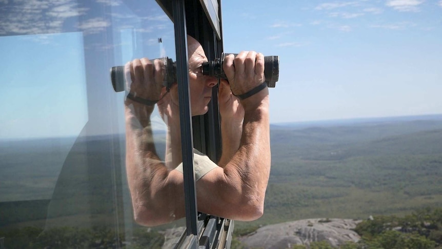A man holding a pair of binoculars looks out over the landscape from a tower.