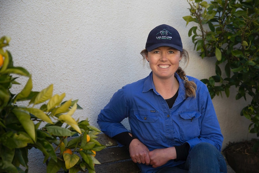 A smiling young woman with a plait, wearing blue work clothes and a cap, sits between some citrus trees.