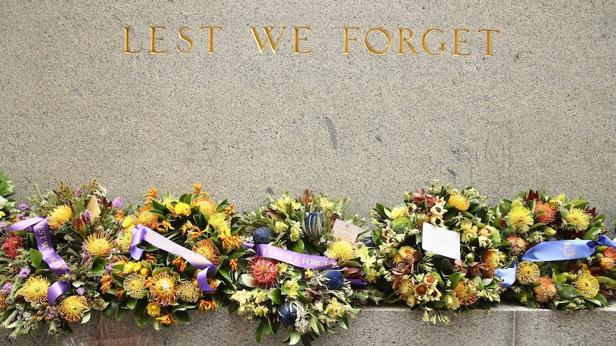 'Lest we forget' lettering on stone wall monument, with flower wreaths sitting below.
