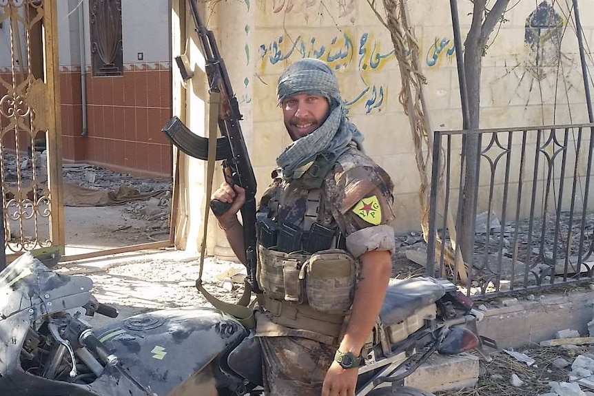 Jamie Williams sits on a motorbike and holds up a gun while smiling.