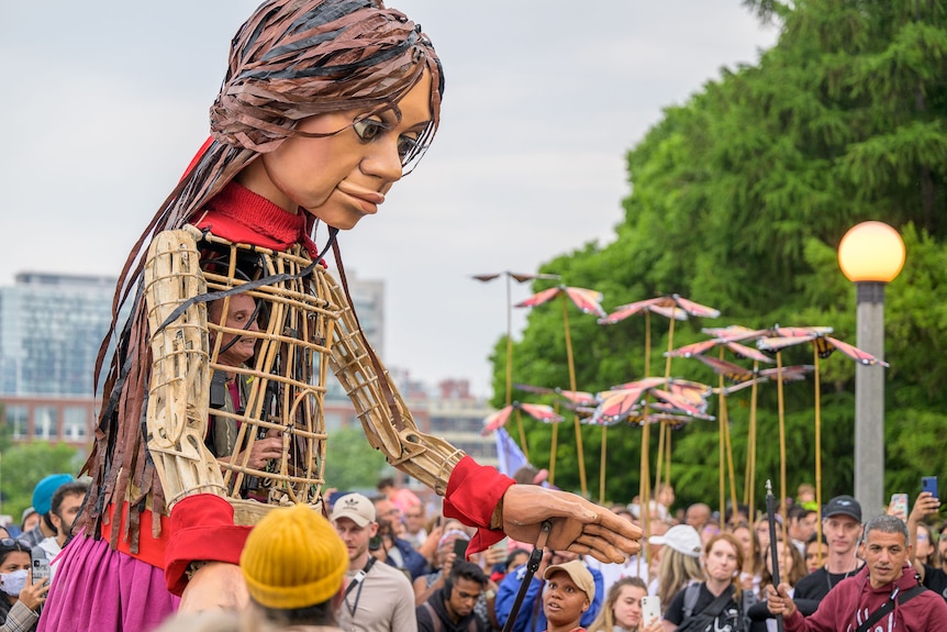 A giant puppet of a girl, made of wicker, with a man inside, leans over a crowd of people in the street.