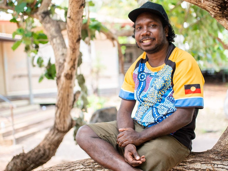 Young man in yellow tee and black cap smiling, sitting on branch of a tree, with building in background.