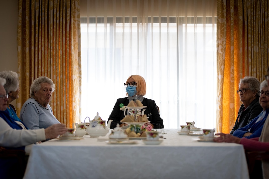 A group of elderly lady sit around a table with tea cups and sandwiches with a woman wearing a mask.