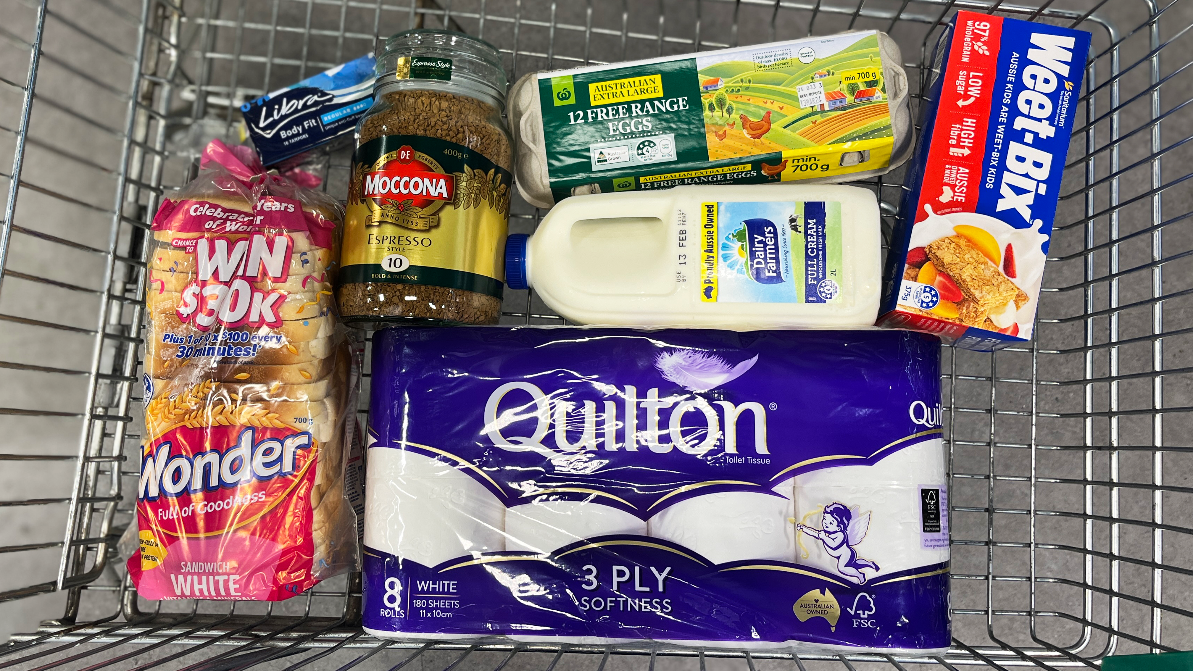 A trolley of grocery store items, including toliet paper, tampons, Weet-bix, white sliced bread, instant coffee, eggs and milk. 