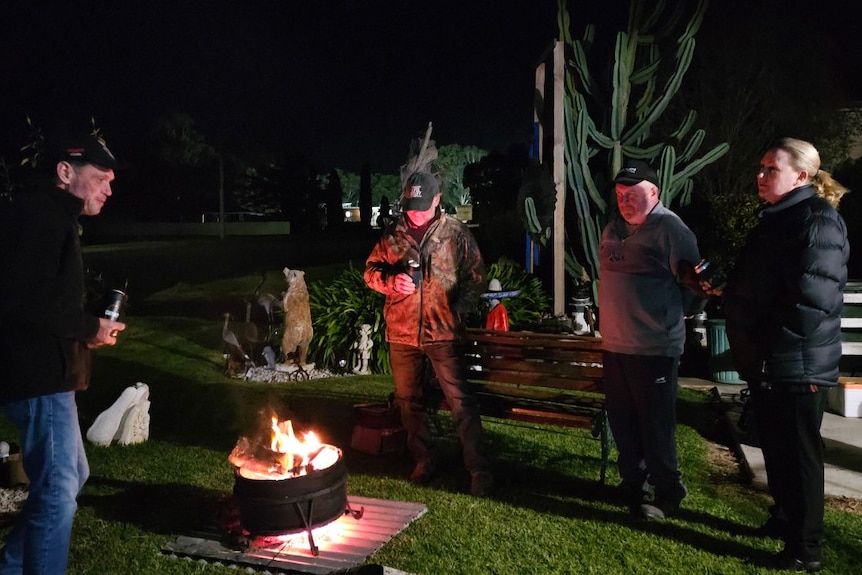 A man and three friends stand around a fire at night in a backyard holding beers. 