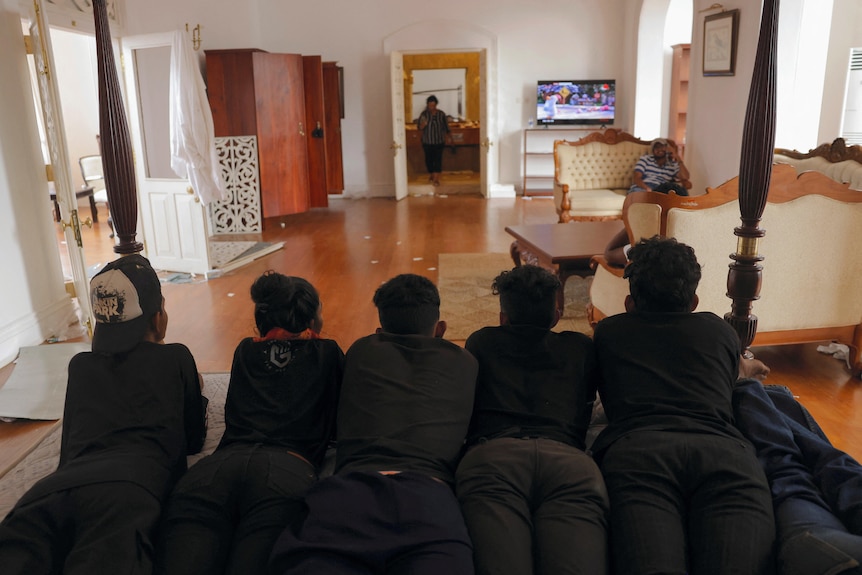 Men dressed in black lay on their stomachs on the president's bed and watch the cricket on a nearby TV. 