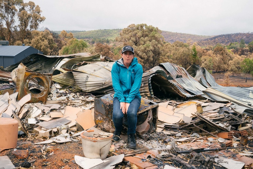 A woman in a blue tracksuit sits on a burnt-out washing machine amongst the wreckage of a bushfire-hit home.