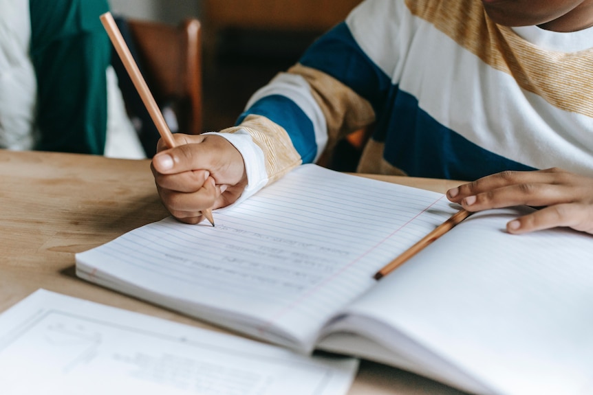 A close up of the hand of a child with brown skin and a striped long sleeve top, holding a pencil and writing in a book.