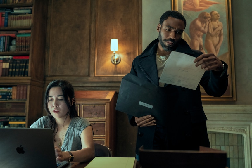 Maya Erskine sitting at a computer and Donald Glover standing holding and looking at some paper in character as Mr & Mrs Smith
