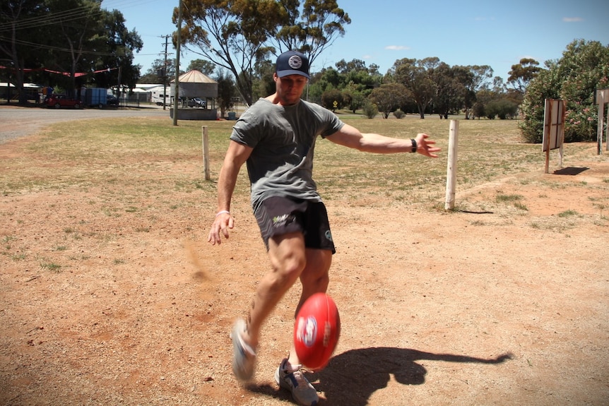 A young man in shorts, t-shirt and hat kicks an Australian Rules Football.