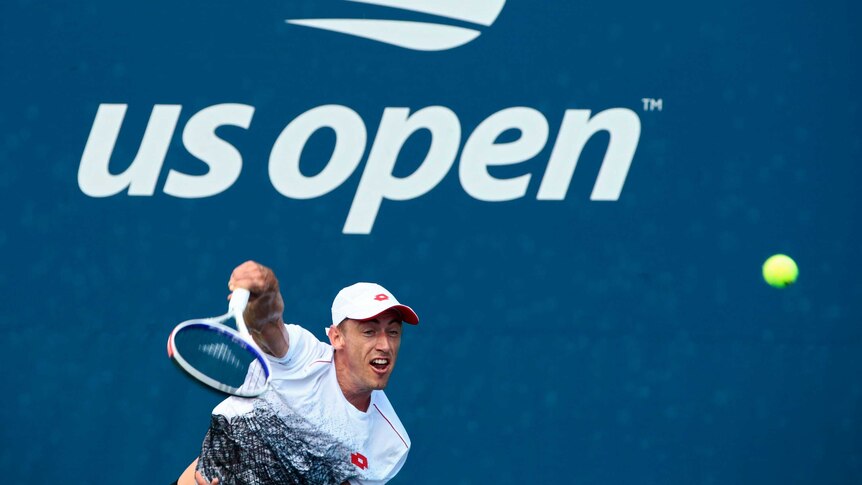 A tennis player in a white shirt and cap serves with a big US Open banner behind him.