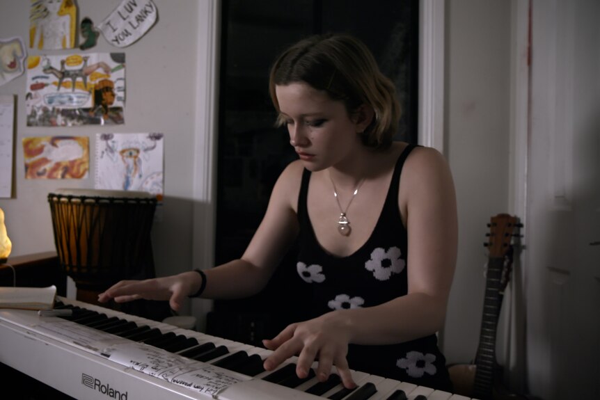 A teenager playing a keyboard in a black top and gold neckless with drawings stuck to the wall behind her