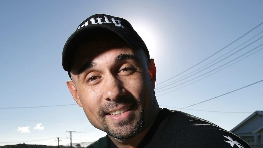 A man wearing a black t-shirt and black cap, holding a pizza box, smiles and points his hand outwards.