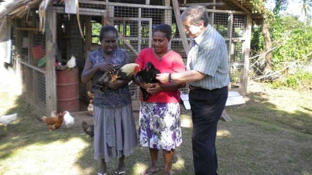Dr Phil Glatz with two local women in Papua New Guinea who are holding chickens in front of a chicken coop.